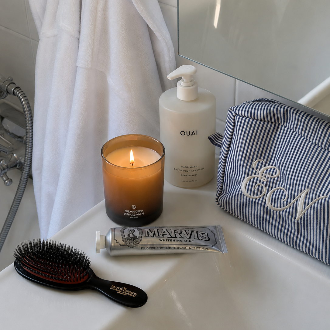 A styled bathroom scene with a lit amber candle, a white pump bottle labeled OUAI, a striped blue and white toiletry pouch with white script embroidery, a tube of MARVIS hand cream, a black hairbrush, and a white bath towel draped over a tiled wall.