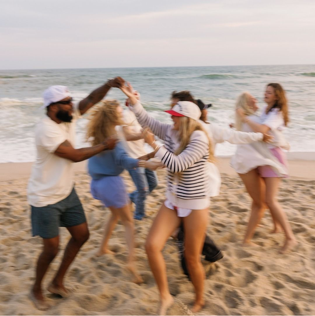 A group of five friends dancing joyfully on a sandy beach, with the ocean and sky in the background. The group is wearing casual summer clothing, including a striped long-sleeve shirt, white t-shirt, denim shorts, and a white cap.