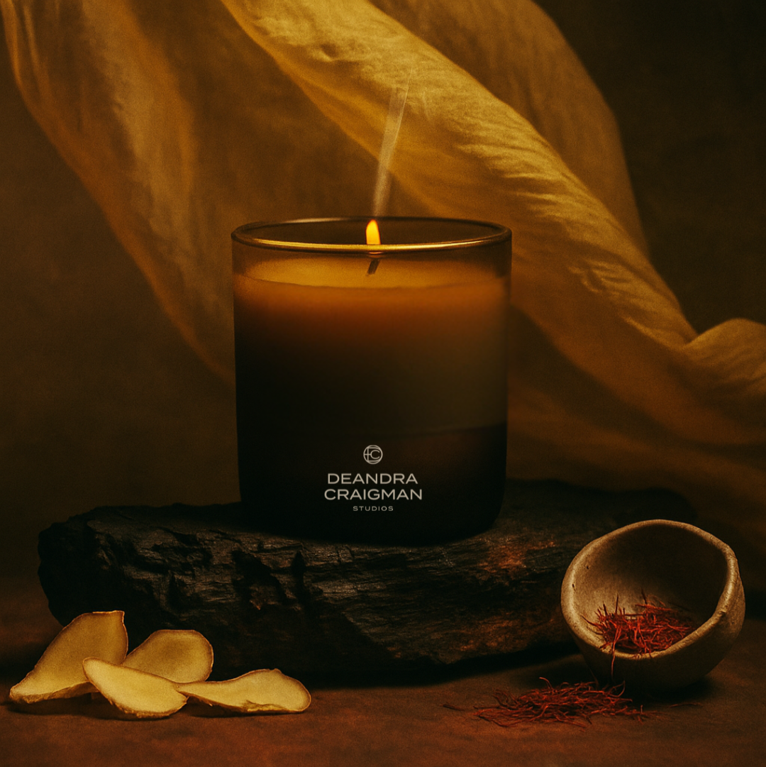Candle with 'DeAndra Craigman Studios' branding on a dark surface with a warm glow.