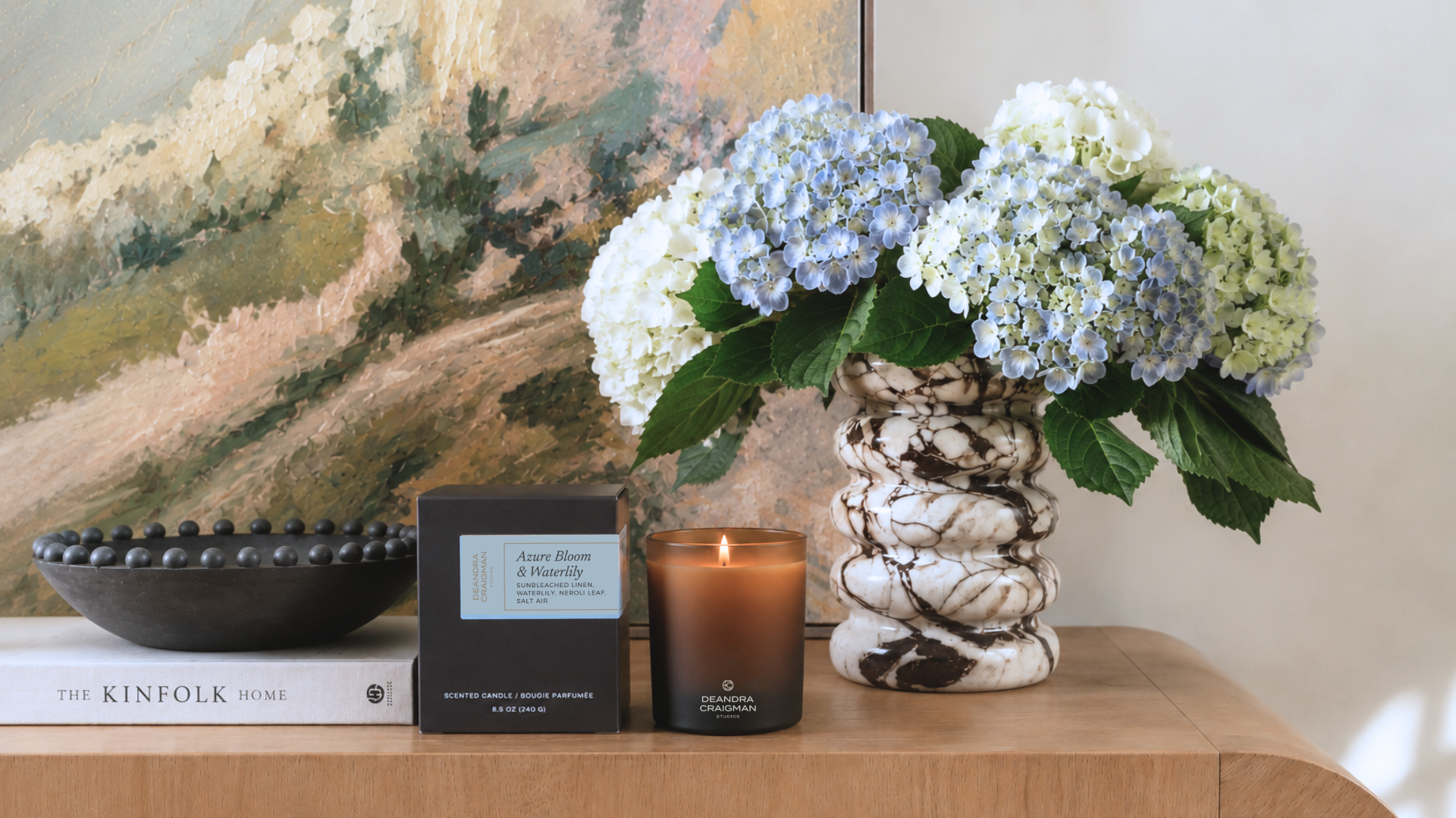 A styled interior scene showing a lit amber glass candle next to a black box and book, with a ceramic vase of blue and white hydrangeas in front of a landscape painting.