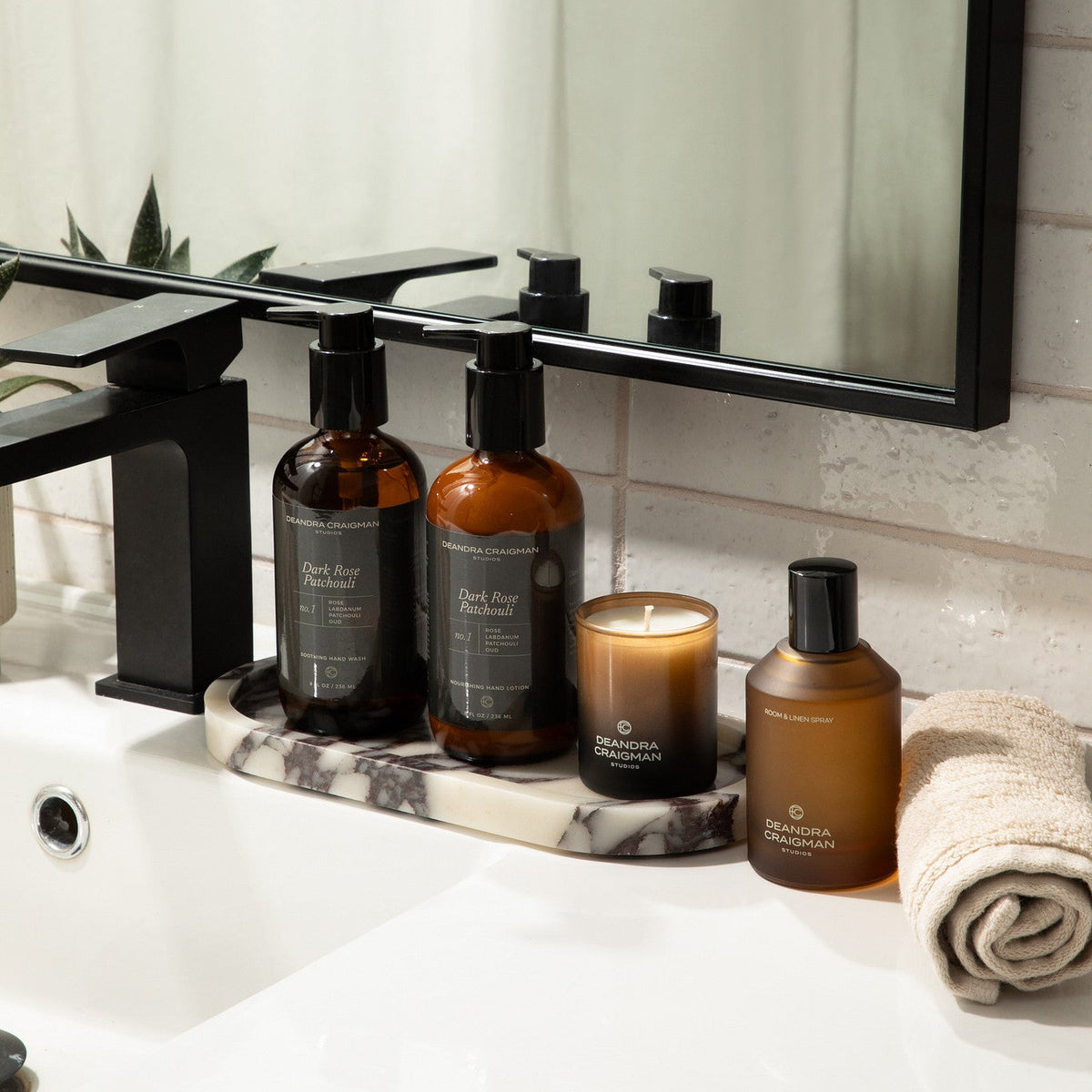 A bathroom vanity scene with several amber glass toiletry bottles and a candle arranged on a marble tray beside a sink and black faucet. 