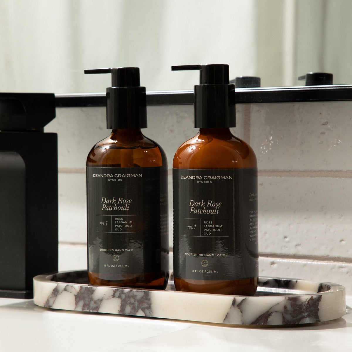 Two amber pump bottles of DeAndra Craigman Studios Dark Rose Patchouli Hand Wash resting on a marble tray on a bathroom counter.