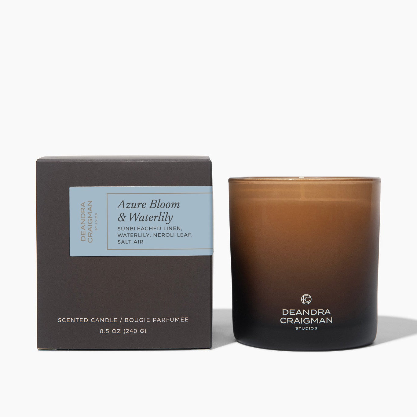 A Deandra Craigman scented candle in an amber glass jar next to its dark brown packaging box, labeled 'Azure Bloom & Waterlily'.